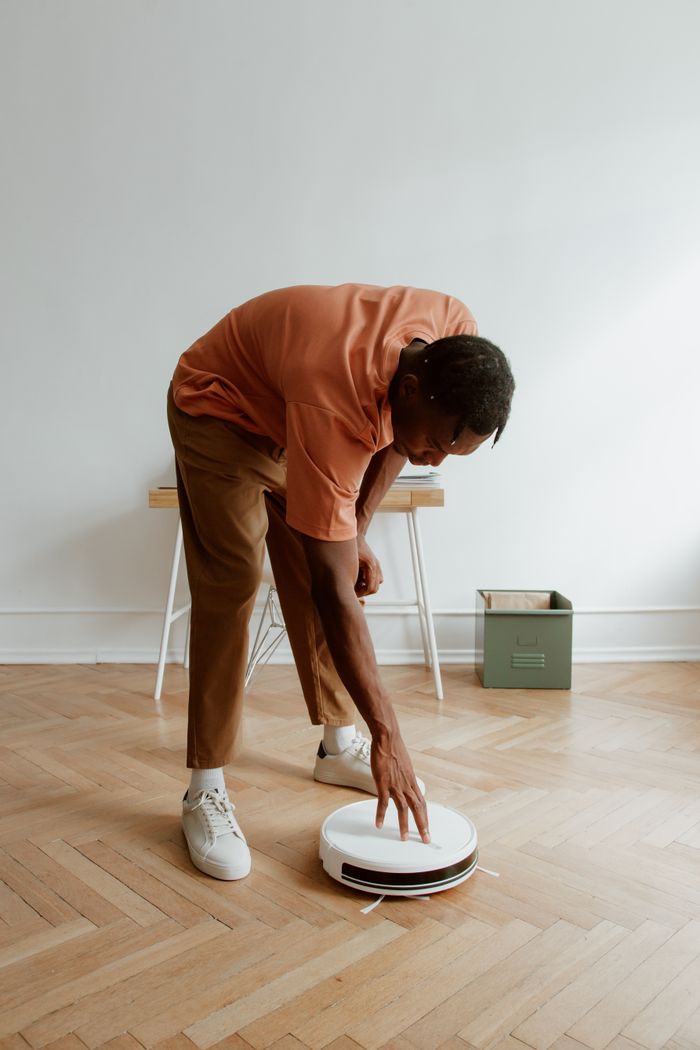 A man operates a robot vacuum cleaner on a wooden floor in a minimalist home setting.
